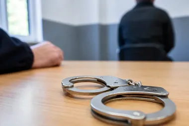 Kajdanki leżą na drewnianym stole w pomieszczeniu, przy którym siedzą dwie osoby w ciemnych ubraniach.